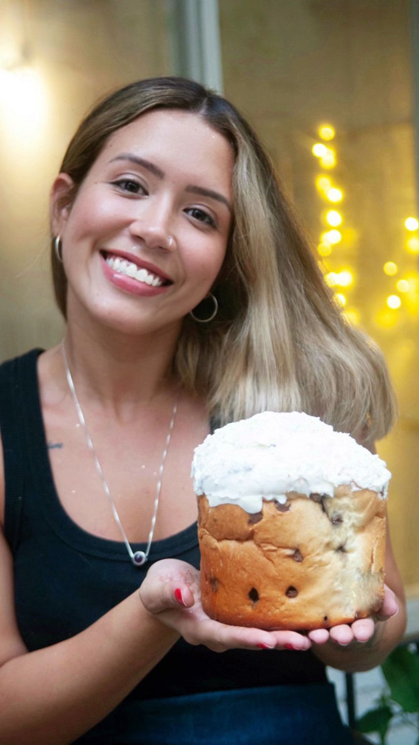 Mulher segurando um panetone com cobertura branca em um ambiente iluminado por luzes decorativas ao fundo.