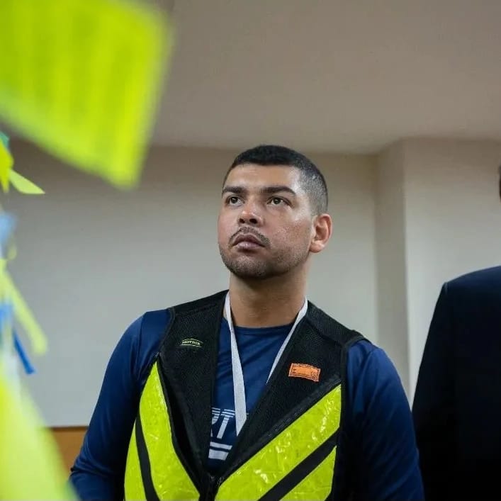 Homem usando colete refletivo amarelo em ambiente interno, olhando para cima com expressão séria.