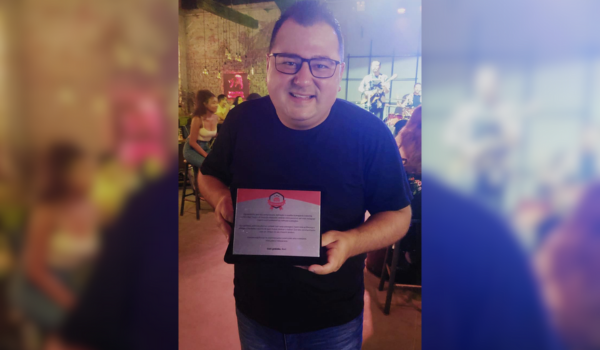 Homem sorridente segurando uma placa comemorativa em um ambiente descontraído, com iluminação baixa e uma banda ao fundo.