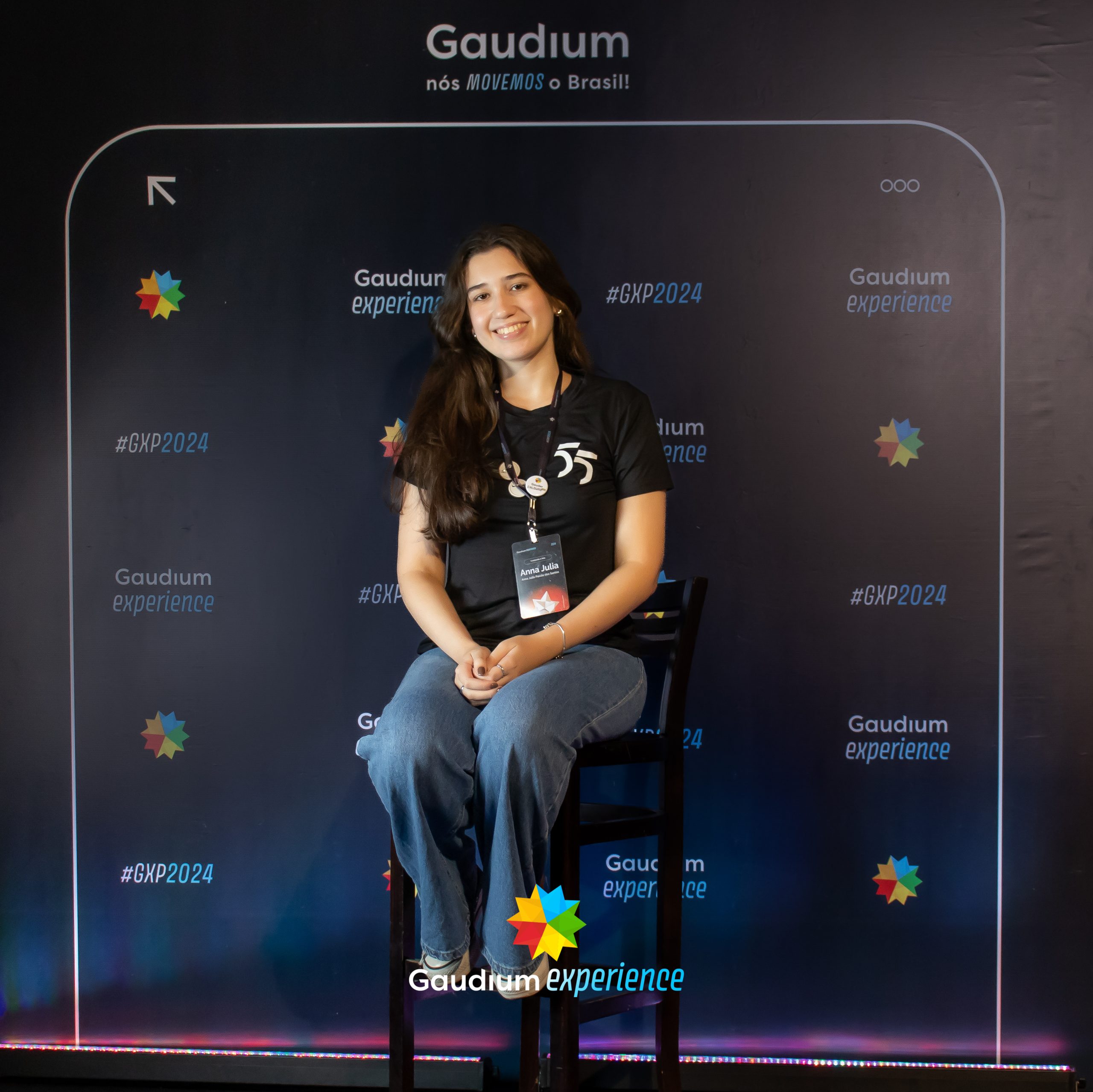 A imagem mostra uma pessoa sentada em uma cadeira em frente a um painel com o tema "Gaudium experience #GXP2024." O painel inclui o logotipo da Gaudium e o slogan "nós MOVEMOS o Brasil!" A pessoa está usando uma camiseta preta com o número "55" estampado, calça jeans e um crachá de identificação com o nome "Anna Júlia." O logo colorido da Gaudium aparece na parte inferior da imagem.