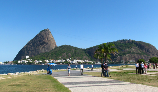 Vista do Aterro do Flamengo, no Rio de Janeiro, com o Pão de Açúcar ao fundo, pessoas caminhando, pedalando e apreciando o espaço ao ar livre.