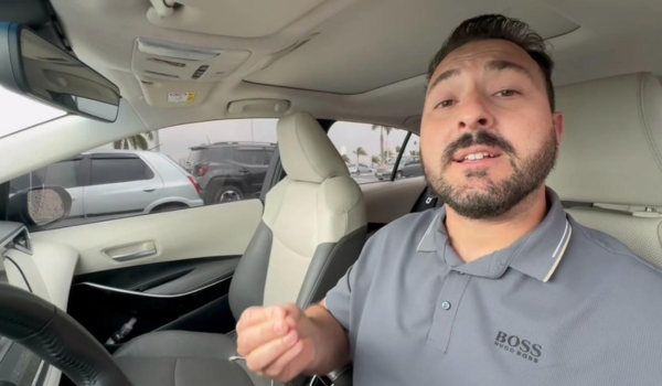 Homem em um carro moderno, vestindo uma camisa polo cinza da Hugo Boss, falando e gesticulando com a mão enquanto está sentado no banco do motorista.