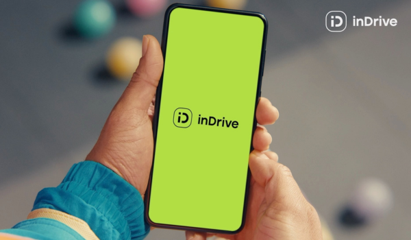 Mãos segurando um smartphone com a tela verde exibindo o logotipo do aplicativo inDrive, em um fundo desfocado com bolas coloridas ao fundo.