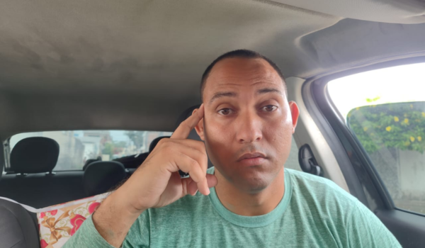 Homem dentro de um carro com expressão séria, encostando a mão no rosto e olhando diretamente para a câmera.