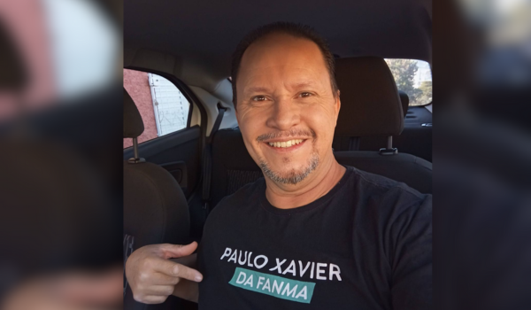 Homem sorridente dentro de um carro, vestindo uma camiseta preta com os dizeres "Paulo Xavier da Fanma".