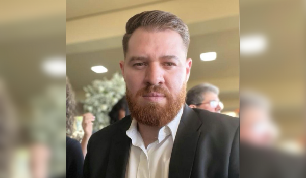 Homem com barba ruiva, vestido com terno preto e camisa branca, em ambiente interno iluminado.