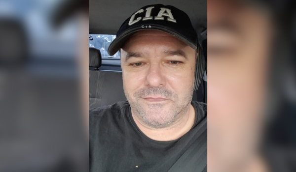 Homem com boné preto escrito "CIA" sentado em um carro, olhando para a câmera.