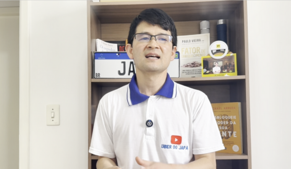 Homem de óculos, vestindo uma camiseta branca com a logo "Uber do Japa" e gola azul, está em frente a uma estante com livros e objetos decorativos, falando ou apresentando algo.