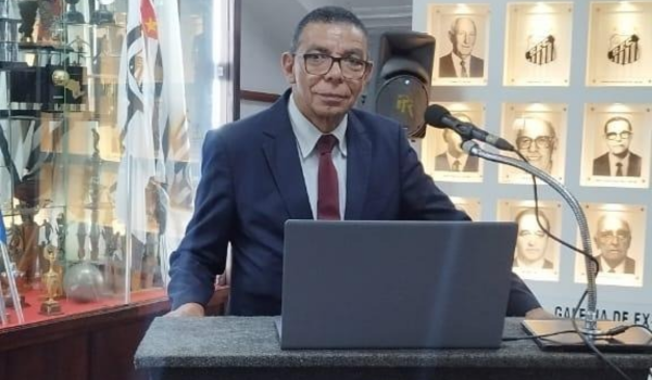 Homem de terno azul-marinho e gravata bordô, com óculos, falando em um púlpito com microfone e laptop, ao fundo há uma parede com fotos de personalidades e uma estante com troféus.