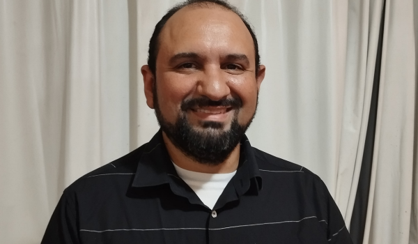 Homem sorridente de barba e cabelo curto, vestindo camisa preta com listras finas brancas, posando em frente a uma cortina branca.