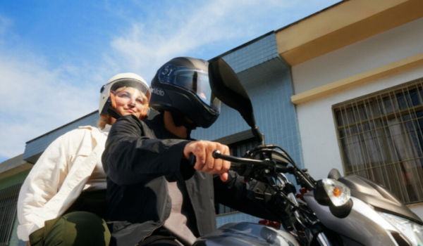 Dois passageiros com capacetes estão em uma moto, sendo conduzidos em uma área residencial.