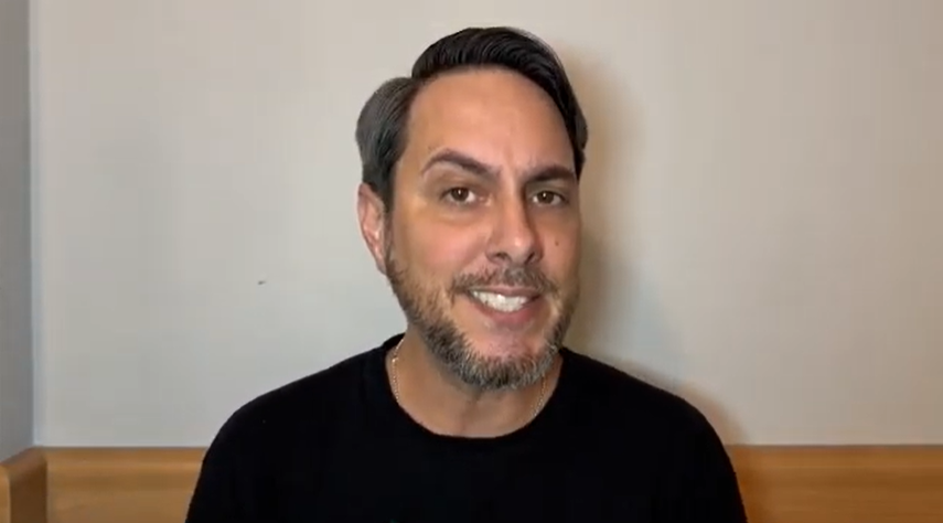 Homem com barba, cabelos curtos e grisalhos, vestido com uma camiseta preta, sentado em frente a uma parede branca.