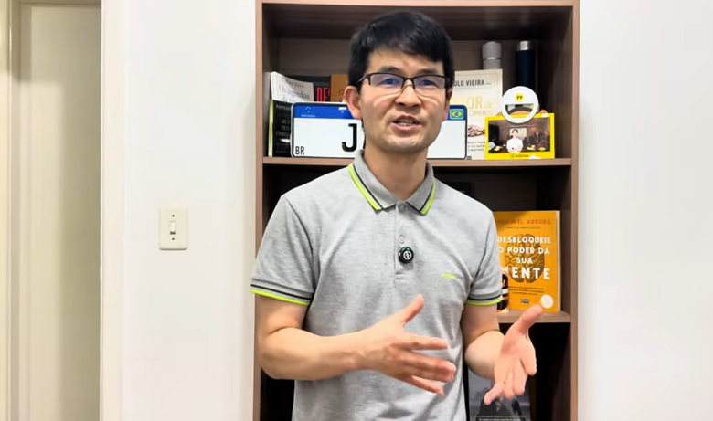 Homem de óculos, usando camisa cinza com detalhes verdes, gesticulando em frente a uma estante com livros e itens decorativos.