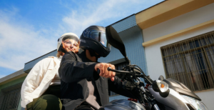 Dois passageiros com capacetes estão em uma moto, sendo conduzidos em uma área residencial.