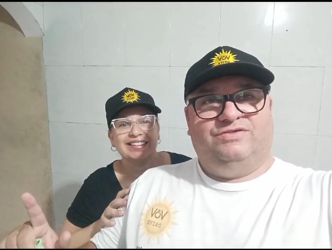 Duas pessoas usando bonés pretos com o logo da V&V Drive, sorrindo para a câmera em um ambiente interno.