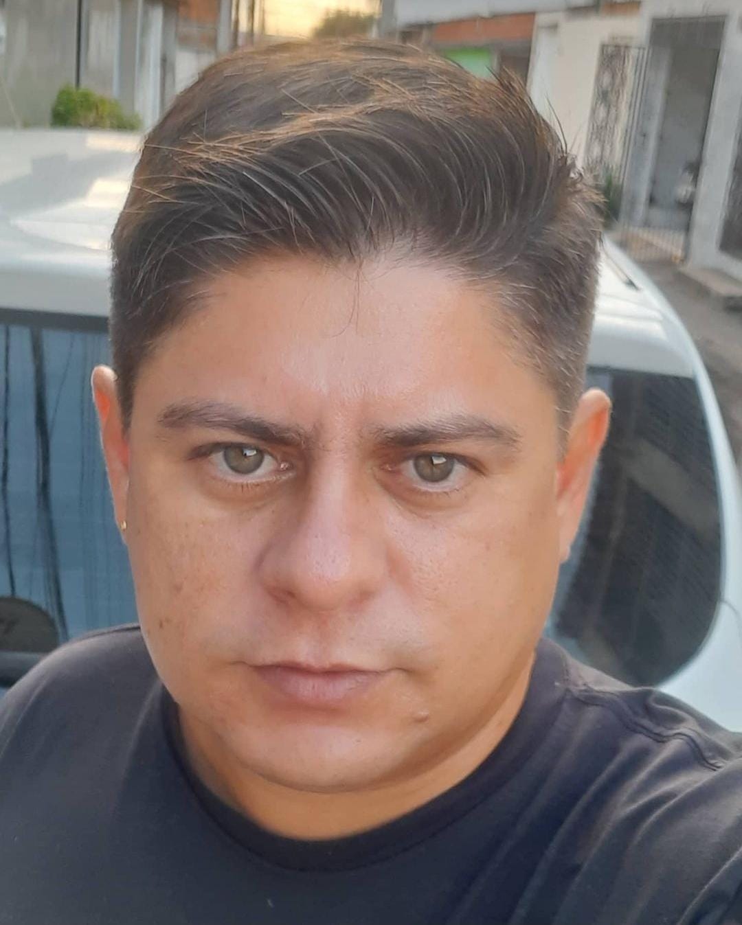 Homem com expressão séria olhando diretamente para a câmera, com cabelos curtos e penteados, em um ambiente ao ar livre com casas e um carro ao fundo.