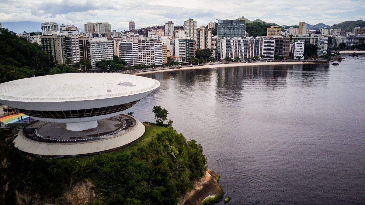 Vista aérea do Museu de Arte Contemporânea (MAC) de Niterói, projetado pelo arquiteto Oscar Niemeyer, localizado no topo de um penhasco à beira-mar. Ao fundo, a praia de Icaraí, em Niterói, com edifícios residenciais ao longo da orla e montanhas ao horizonte.