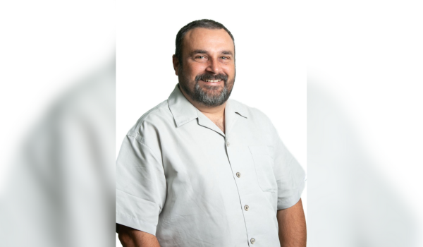 Homem sorridente com barba e cabelos curtos, vestindo uma camisa social clara de mangas curtas, posando contra um fundo branco.