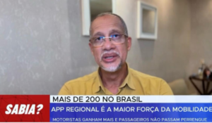 Homem de óculos e barba grisalha veste camisa amarela e fala diante de uma câmera, com um fundo neutro de parede clara e quadros. A imagem contém uma faixa informativa destacando o crescimento dos aplicativos regionais de mobilidade no Brasil.