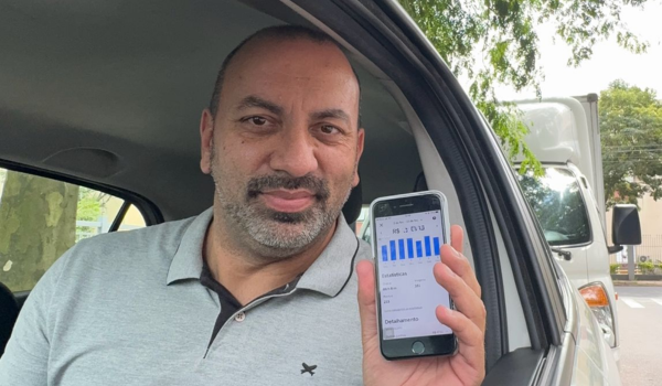 Homem dirigindo um carro, usando óculos escuros e camisa azul. A imagem é capturada do ponto de vista do passageiro, com o volante em primeiro plano e a paisagem verde ao fundo.