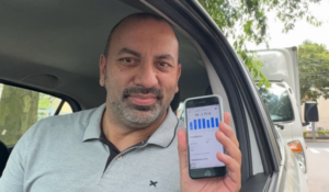 Homem dentro de um carro segura um smartphone exibindo um gráfico na tela, sorrindo levemente para a câmera.