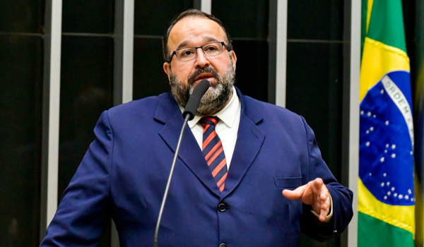 Homem de terno azul e gravata listrada vermelha e preta discursa em um microfone, gesticulando com a mão direita. Atrás dele, há uma bandeira do Brasil.