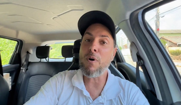 Motorista de aplicativo dentro de um carro, usando boné preto e camisa branca, falando diretamente para a câmera com expressão animada. O fundo mostra o interior do veículo e uma rua com casas ao fundo.