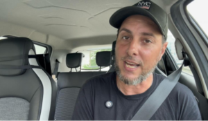 Homem de barba curta e cabelo escuro usando boné preto e camiseta preta, sentado no banco do motorista de um carro. Ele está usando cinto de segurança e parece estar falando ou gravando um vídeo.