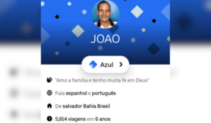 Captura de tela de um perfil em um aplicativo de transporte. A imagem mostra um motorista chamado João com um fundo azul decorado com formas geométricas. Abaixo da foto de perfil, há informações como idioma falado (espanhol e português), local de origem (Salvador, Bahia, Brasil), e o total de viagens realizadas (5.914 em 6 anos). Também há uma seção de elogios recebidos com diferentes ícones e números.