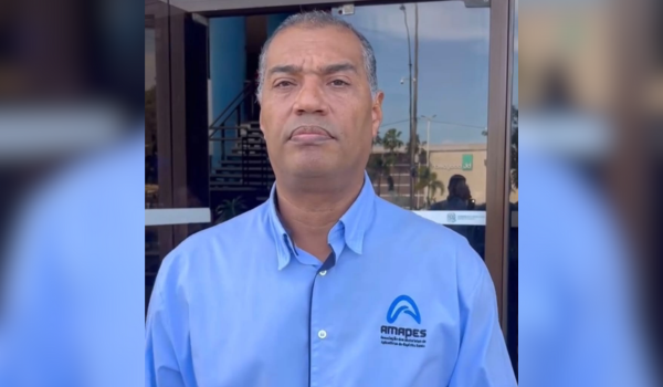 Homem de meia-idade com cabelo curto e grisalho, vestindo uma camisa azul clara com o logotipo da AMAPES, posando em frente a um edifício comercial de vidro.