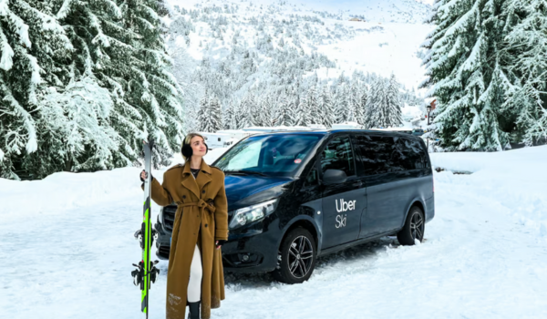 Mulher de sobretudo marrom segura um par de esquis ao lado de uma van preta com a logo "Uber Ski", estacionada em uma estrada coberta de neve, cercada por árvores e montanhas nevadas ao fundo.