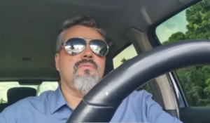 Homem dirigindo um carro, usando óculos escuros e camisa azul. A imagem é capturada do ponto de vista do passageiro, com o volante em primeiro plano e a paisagem verde ao fundo.