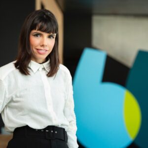 Mulher de cabelos castanhos e franja, vestindo camisa branca e calça preta, sorrindo para a câmera. Ao fundo, há um elemento decorativo azul e verde em um ambiente corporativo.