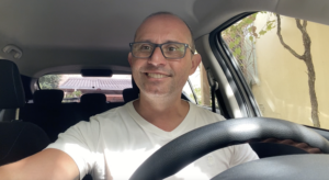 Homem sorridente dirigindo um carro, usando óculos e camisa branca.