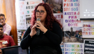 Mulher de cabelos cacheados e tingidos de vermelho, usando óculos e roupa preta, fala ao microfone em um ambiente interno decorado com cartazes e faixas contendo frases e reivindicações.