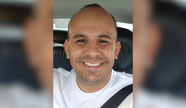 Homem sorridente usando camiseta branca, sentado no banco do motorista de um carro, com cinto de segurança afivelado.