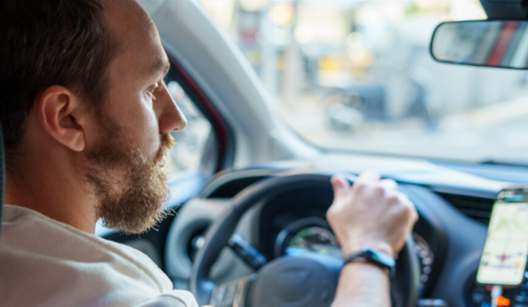 Homem dirigindo um carro, visto de perfil, com atenção à rua enquanto segura o volante com uma das mãos. Um celular com aplicativo de navegação está fixado no painel.