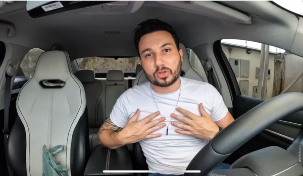 Homem com barba e cabelo curto, sentado no banco do motorista de um carro, usando uma camiseta branca e um colar. Ele gesticula com as mãos sobre o peito enquanto fala, olhando para a câmera.