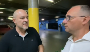 Dois homens conversam em um estacionamento coberto, um usando camisa preta e o outro de camisa branca com óculos.