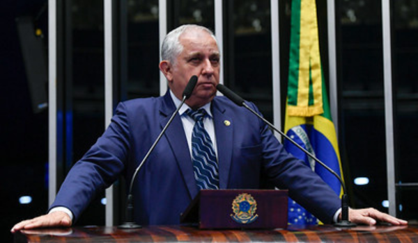 Homem de terno azul fala ao microfone em plenário com a bandeira do Brasil ao fundo.