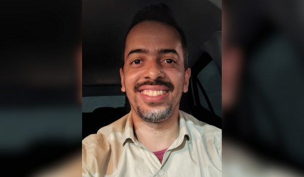 Homem sorridente dentro de um carro, vestindo uma camisa bege e com barba por fazer.