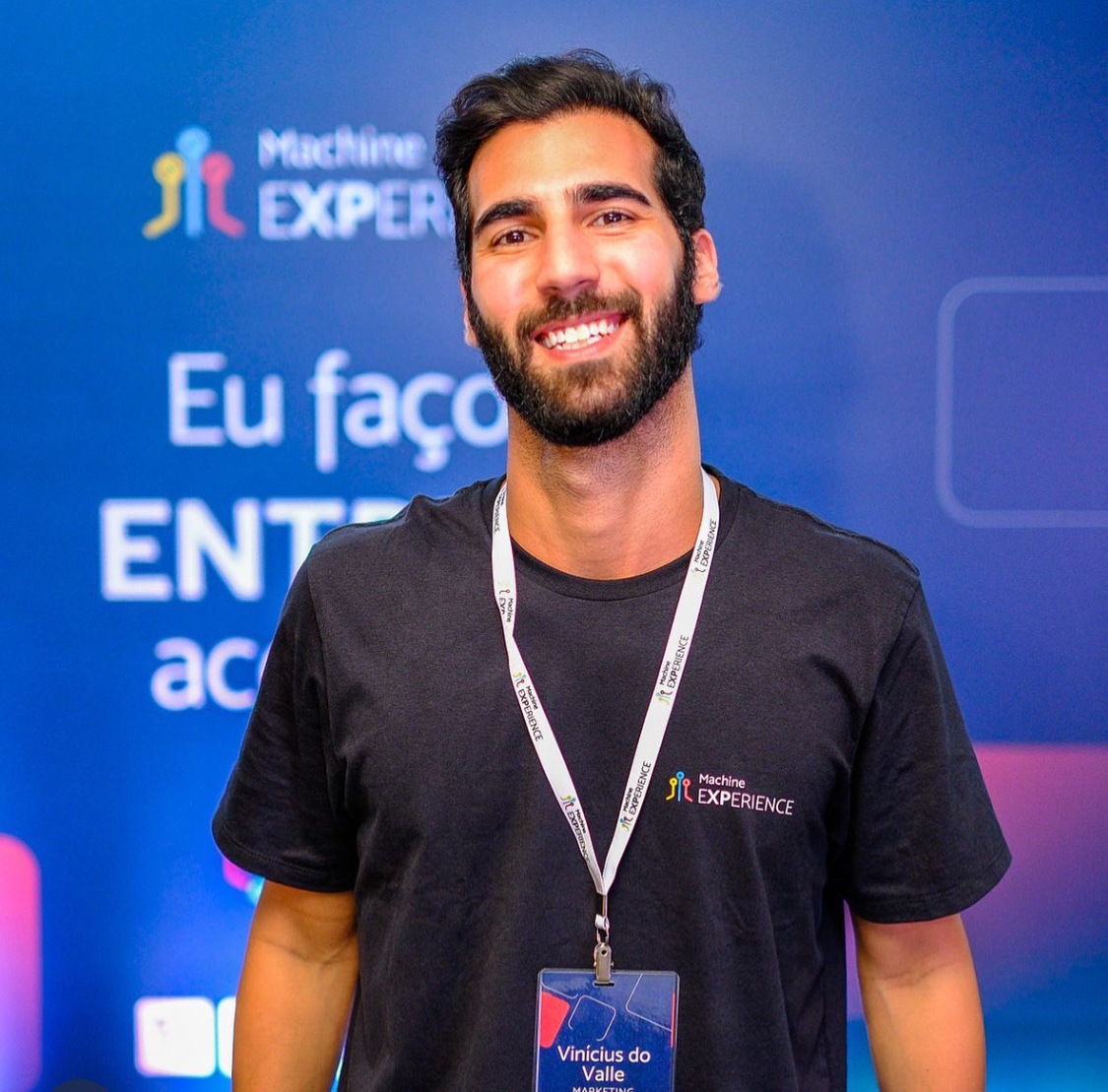 Vinícius do Valle sorrindo no evento Machine Experience, vestindo uma camiseta preta com o logo do evento e um crachá.