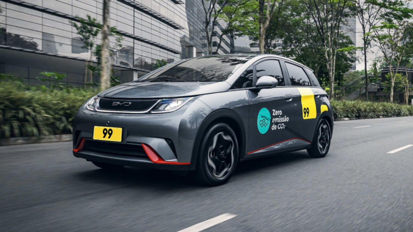 Carro elétrico da empresa 99 com adesivo “Zero emissão de CO?” circulando em via urbana arborizada, com prédios modernos ao fundo.