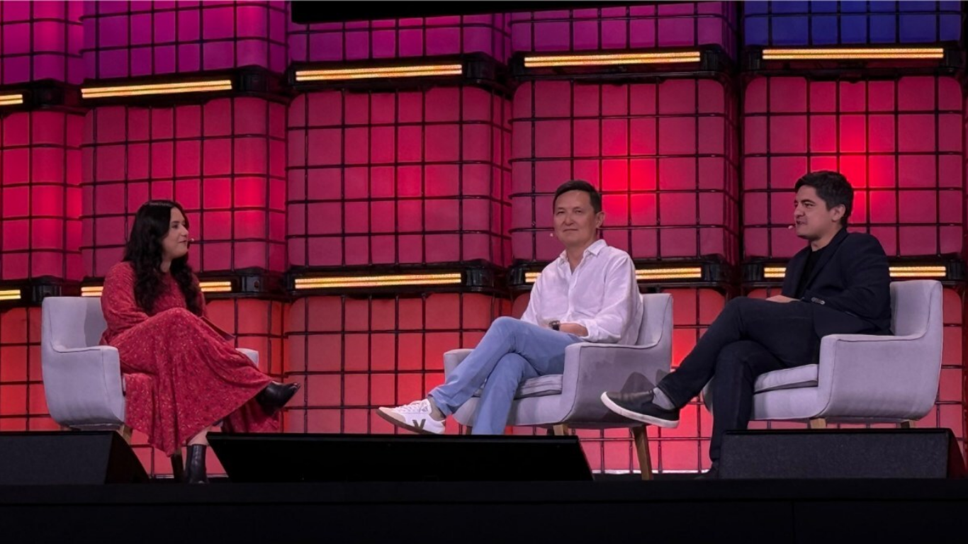 Três pessoas sentadas em poltronas em um palco iluminado com fundo rosa e lilás durante um painel ou talk show.