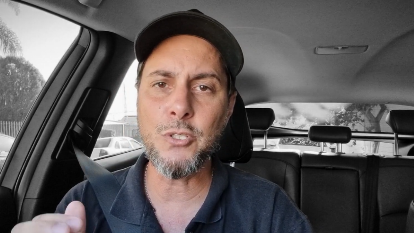Homem de boné preto e barba grisalha falando dentro de um carro, com o cinto de segurança afivelado.