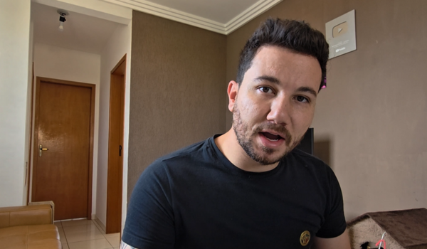 Homem de cabelo curto e barba rala, usando camiseta preta, olhando diretamente para a câmera em um ambiente interno com paredes marrons e portas de madeira ao fundo.