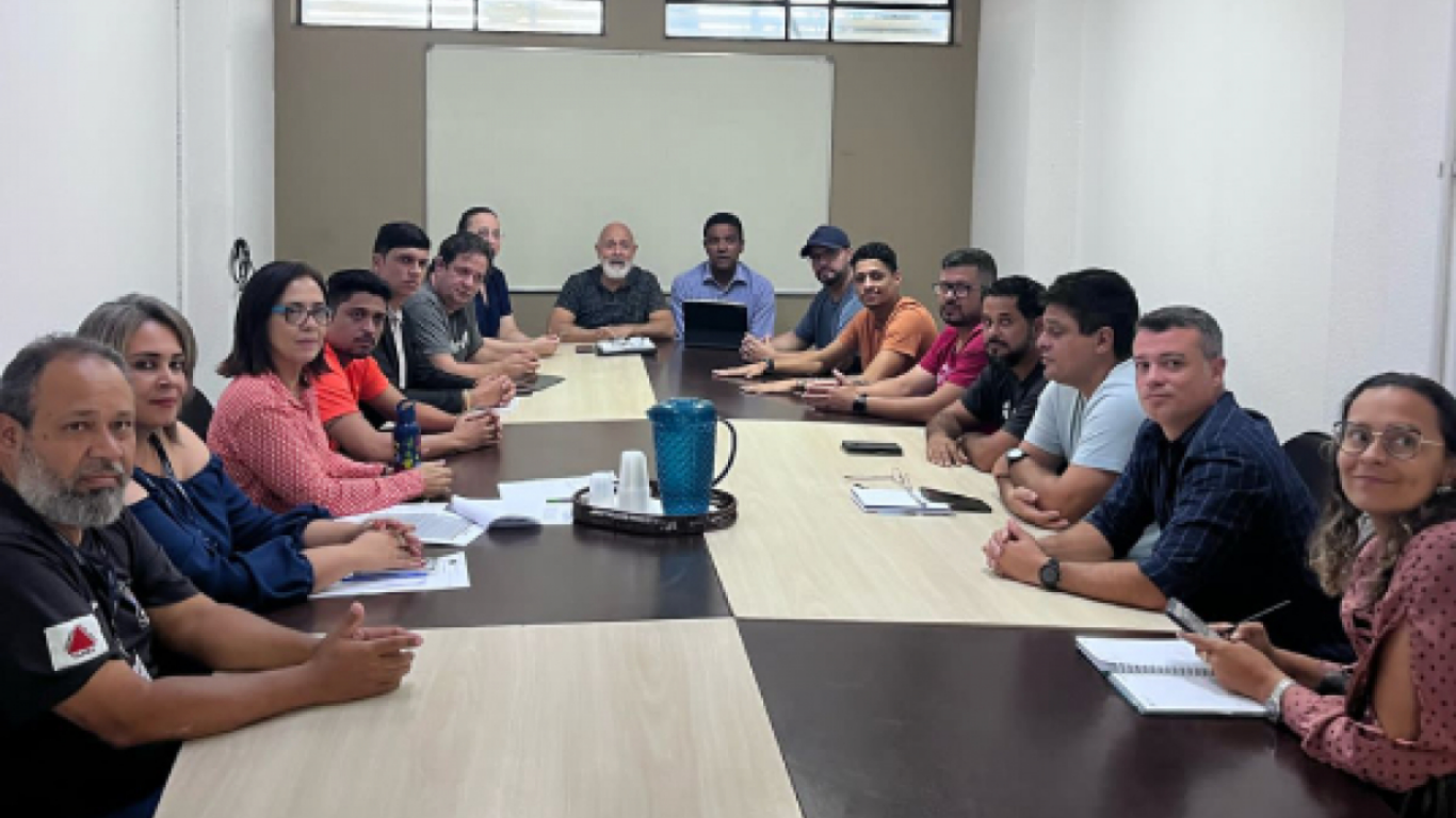 Reunião com cerca de 18 pessoas sentadas em uma longa mesa retangular em uma sala de reuniões, todas voltadas para a câmera.