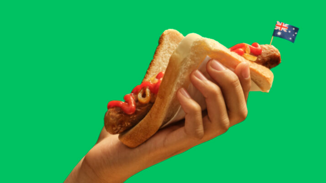 Mão segurando um pão de forma com salsicha, ketchup e mostarda, enfeitado com uma pequena bandeira da Austrália.