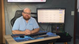 Homem com barba e óculos sentado à mesa de escritório com computador e duas telas, vestindo camisa azul clara com logo da iMOVE.