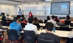 Auditório com pessoas sentadas assistindo a uma apresentação de duas pessoas em frente a um projetor.